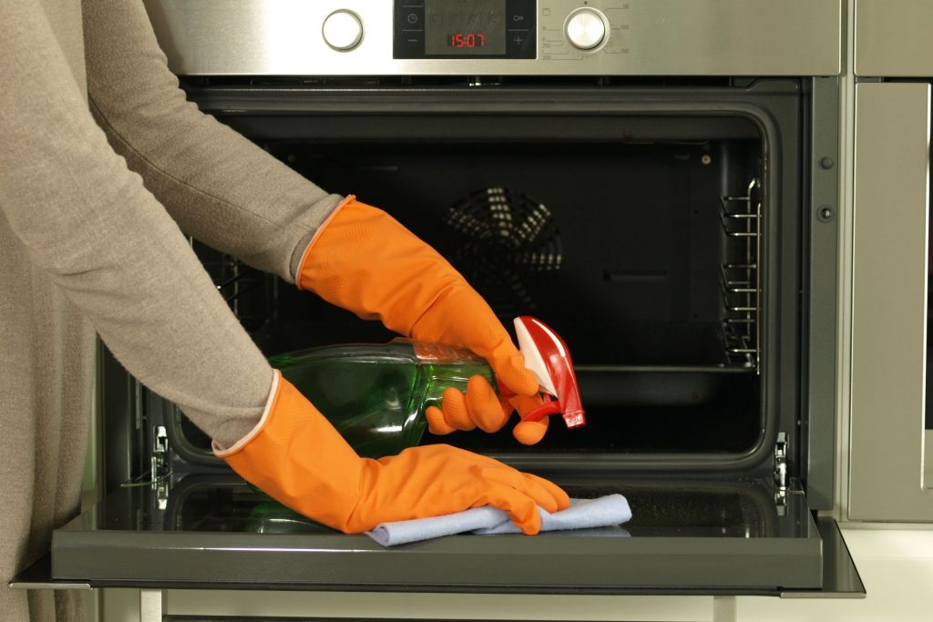 A woman wearing orange gloves uses an oven cleaner on the inside of her appliance. 